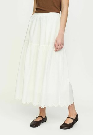 Woman wearing a white ankle-length skirt with scalloped hem and eyelet details, paired with a brown sleeveless top and black Mary Jane shoes.