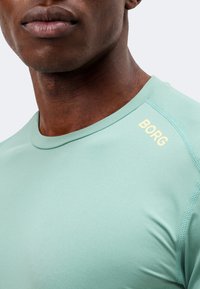 Κοντινό πλάνο ενός ατόμου που φοράει ανοιχτοπράσινο πουκάμισο με την επιγραφή "BORG" σε ωχρό κίτρινο χρώμα στον δεξιό ώμο.