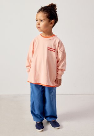 Jedes kleines Kind mit lockigem Haar, zu einem Dutt gebunden, trägt einen übergroßen pfirsichfarbenen Pullover, blaue Hosen und marineblaue Slipper, steht und schaut nach links.