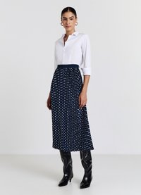 Chemise blanche à col, associée à une jupe plissée bleu marine à pois blancs, complétée par des bottes noires à talons montant jusqu'aux genoux.