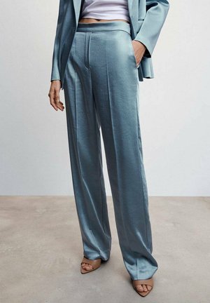 Person trägt hellblaue Satinhose mit weitem Bein, passenden Blazer, weißes kurzes Top und beige offene Schuhe, steht auf grauem Boden.