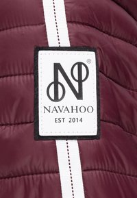 Giacca puffer bordeaux con cuciture bianche e un patch rettangolare con un "N" intrecciato e un accento circolare, etichettato "NAVAHOO EST 2014."