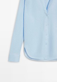 Chemise bleu clair à manches longues avec patte de boutonnage, ourlet arrondi et poignets à un bouton sur fond blanc.