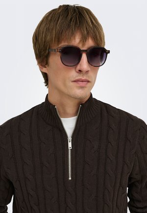 Homme portant des lunettes de soleil rondes foncées et un pull en maille torsadée noir avec un col à demi-fermeture éclair par-dessus une chemise blanche.