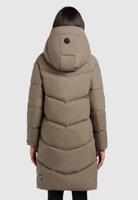 Persoon in een lange, gewatteerde taupe jas met capuchon, zichtbare stiksels en een klein zwart logopatch nabij de zoom, met de rug naar de kijker gekeerd.