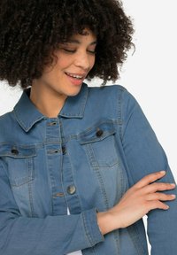 Veste en denim bleu clair, avec un col, des boutons-pression à l'avant, deux poches poitrine et des coutures contrastées. Texture lisse.