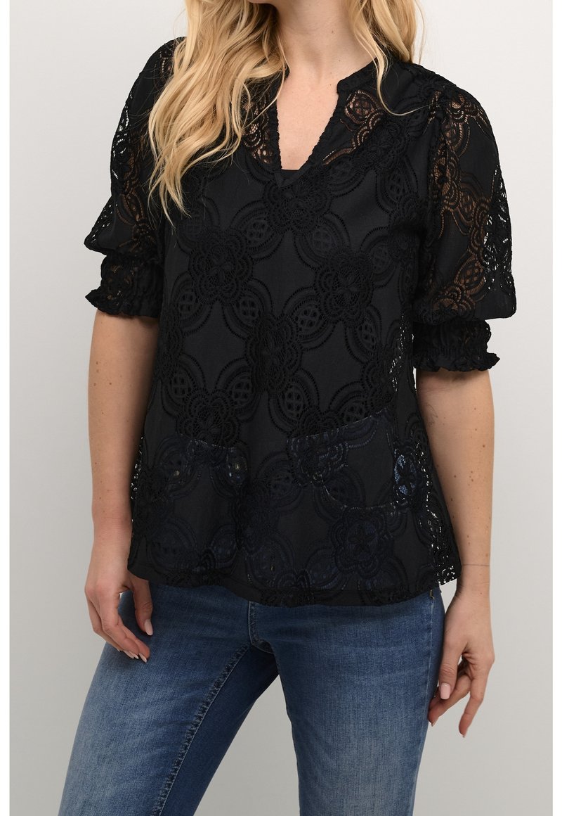 Femme portant un chemisier noir à motif dentelle avec manches trois quarts et un jean bleu, debout sur un fond uni.