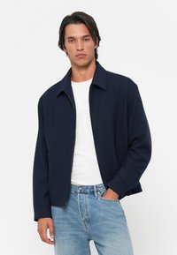 Mornarska zip-up jakna s razširjenim ovratnikom, dolgimi rokavi in stranskimi žepi, izdelana iz gladkega materiala. Obuta čez belo majico in svetle jeans hlače.