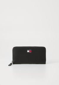 Svart glidelås-lommebok i glat materiale, med rektangulær form og en logoplade med røde, hvite og blå detaljer på forsiden.
