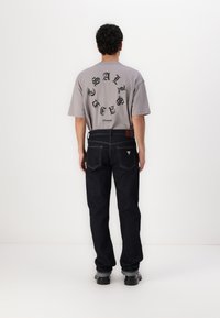 Camiseta gris de gran tamaño con texto gráfico circular negro en la parte posterior, combinada con jeans de mezclilla oscura y calzado negro.