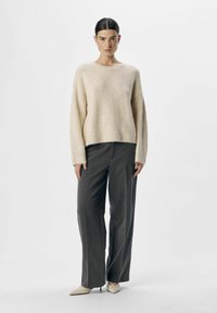 Un pull en tricot beige clair avec des épaules tombantes, associé à un pantalon large gris taille haute et des escarpins beiges à bout pointu.