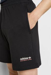 Shorts sportivi neri con cintura elastica e tasche laterali. Presentano il logo "adidas ADVENTURE" in rosa sull'orlo. Tessuto morbido.