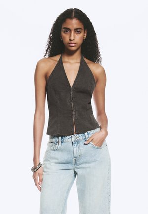 Femme aux cheveux bouclés portant un top dos nu noir et un jean bleu clair, debout avec une main dans la poche et une expression neutre.