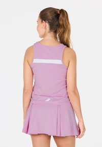 Donna con una coda di cavallo marrone che indossa un top sportivo senza maniche rosa e una gonna plissettata coordinata, in piedi di spalle contro uno sfondo semplice.