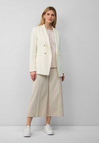 Weißes, doppelt eingeknöpftes Blazer über einer gemusterten cremefarbenen Bluse, kombiniert mit weiten beige Culottes und weißen Sneakers. Sanfte Texturen durchweg.
