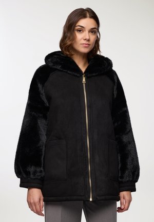 Donna che indossa una giacca nera con cappuccio e maniche e colletto in pelliccia sintetica, in piedi contro uno sfondo chiaro e semplice.