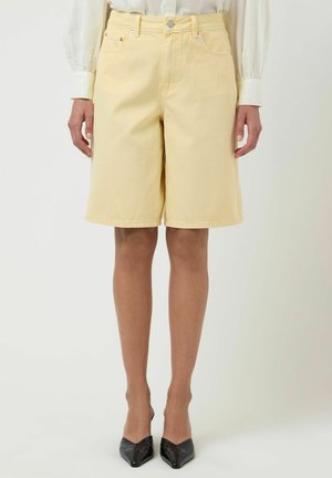 Kvinne som har på seg høytlivede, blekgule denim shorts til knelengde, hvit bluse med pufferm og svarte spisse hæler.