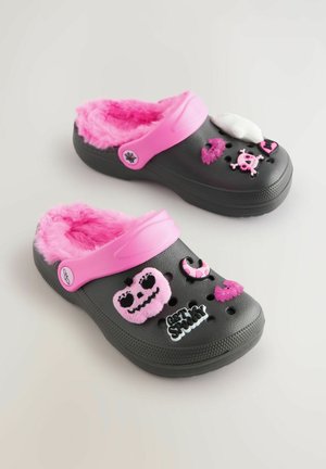 Zuecos negros con forro de piel sintética rosa, que presentan diversos charms decorativos, incluyendo un fantasma, una calavera y el texto "PÓNTETE MIEDO".