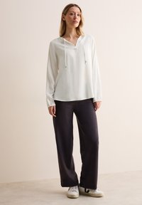 Witte blouse met lange mouwen, een V-hals en een koord, gecombineerd met donkere wijde broek en witte sneakers met zwarte accenten. Soepele texturen.