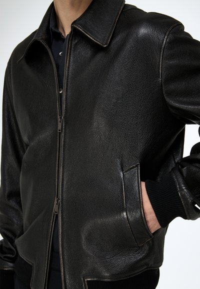 Persona con una chaqueta bomber de cuero negra con cremallera frontal y la mano en el bolsillo lateral, sobre un fondo liso.