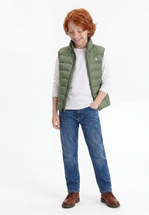 Αγόρι με κόκκινα μαλλιά, που φοράει πράσινο φουσκωτό γιλέκο, λευκό μακρυμάνικο μπλουζάκι, μπλε τζιν και καφέ μπότες, στέκεται με το ένα χέρι στην τσέπη, χαμογελώντας.