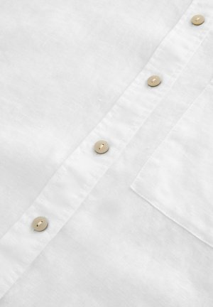 Bílá lněná košile s knoflíkovým zapínáním vpředu a béžovými knoflíky, má také kapsu na levé straně hrudi. Textura látky je hladká s mírně zmačkaným vzhledem.