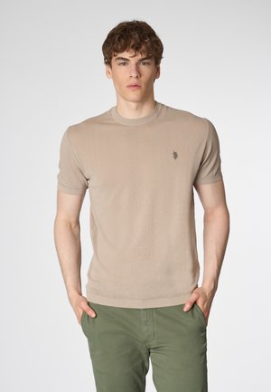 Maglietta a maniche corte in maglia beige con colletto a costine, caratterizzata da un piccolo logo ricamato sul petto; indossata con pantaloni verdi oliva.