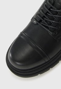 Botte en cuir noir à bout rond, avec un capuchon doublé et des lacets noirs, présentée sur un fond gris clair.