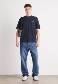 Tricou din bumbac de culoare bleumarin cu un logo mic, asortat cu blugi de denim de culoare albastru deschis și adidași albi. Modelul poartă o geantă crossbody neagră.