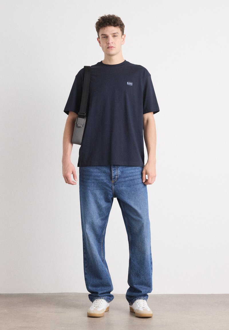 T-shirt in cotone blu navy con un piccolo logo, abbinata a jeans in denim azzurro e sneakers bianche. Il modello porta una borsa a tracolla nera.