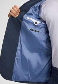 Interno di una giacca blu navy che mette in mostra una fodera azzurro chiaro liscia, una tasca frontale con chiusura a bottone e un'etichetta che recita "MATERIALE ELASTICO FINEST".