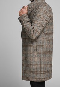 Cappotto a quadri grigio con dettagli marroni, caratterizzato da un colletto strutturato, chiusura con bottoni e tasche laterali, indossato da un modello.