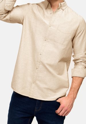 Homme portant une chemise beige à boutons avec les manches retroussées et un jean bleu foncé, une main dans la poche et l'autre levée près du visage.