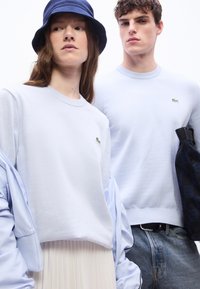 Νεαρή γυναίκα και άντρας που φορούν ανοιχτό μπλε πουλόβερ Lacoste, η γυναίκα με καπέλο τύπου μπαντάνα και πτυχωτή φούστα, ο άντρας κρατά μια τσάντα με καρό σχέδιο.