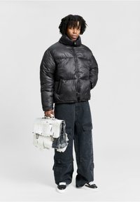 Person iført en sort pufferjakke og løse sorte cargo-bukser, holder en slidt hvid og sort aktentaske, stående foran en ensfarvet baggrund.