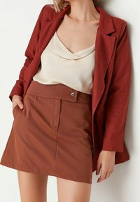 Femme portant un blazer et une jupe couleur rouille avec un haut ivoire sans manches à col bénitier, debout, une main dans la poche, sur un fond uni.