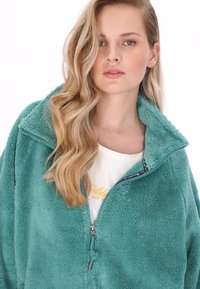 Světlý petrolejový fleeceový pulovr se zipovým límcem, který má měkký, strukturovaný povrch. Nasažený přes bílou košili se žlutým nápisem.