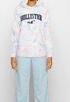 Person iført pastel tie-dye Hollister hættetrøje med forlommer og lyseblå vidde jeans mod en ensfarvet baggrund.