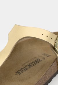 Goldene Leder-Sandalen mit einem dünnen Riemen, verstellbarem Schnallenverschluss und einer konturierten braunen Fußbett. Auffälliges Markenlogo auf der Innensohle gedruckt.