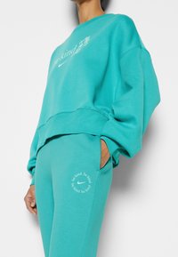Teal cotton sweatshirt with a round neckline, dropped shoulders, and white text. Matching teal joggers feature a circle logo and text detail.