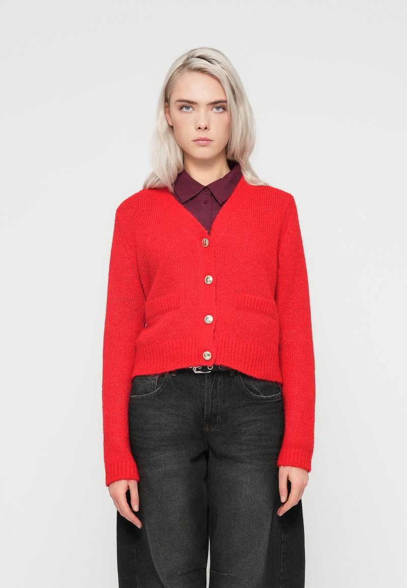 Punane kudumiskardigan V-kaelusega, kahe esisegmendiga taskuga ja viie hõbedase nupuga, kantuna tumeda kraega särk ja mustad püksid.