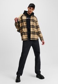 Camisa de franela a cuadros en beige y negro sobre una sudadera negra, combinada con pantalones negros y botas oscuras. El modelo está de pie con un brazo levantado.