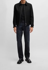 Giacca nera con zip e colletto, indossata sopra una camicia nera, abbinata a jeans in denim scuro e scarpe di pelle nere, in piedi contro uno sfondo grigio.