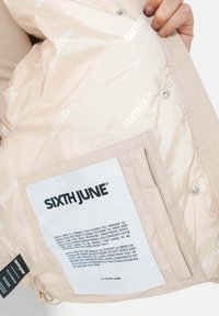 Lichtbeige pufferjack interieur met een glanzende textuur. Heeft een zak met een wit label waarop merkinformatie in zwarte tekst staat.