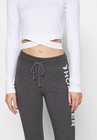 Vit ribbad långärmad crop top med korsdesign vid midjan. Grå sweatpants med dragsko och vit "HO"-text på sidan.