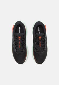 Baskets de sport noires avec un dessus en mesh respirant, accents orange, éléments réfléchissants et une semelle en caoutchouc vert menthe. Comprend un système de laçage.