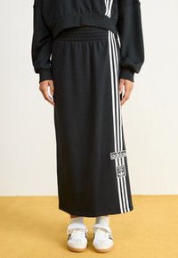 Fekete sportos szoknya fehér függőleges csíkokkal, elasztikus derékpánttal és Adidas logóval. Fehér sportcipővel és fehér zoknikkal van stílusosan párosítva.