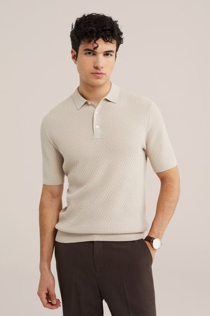 Jeune homme aux cheveux bouclés portant un polo beige texturé à manches courtes et un pantalon marron foncé, debout avec une main dans la poche.