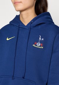 Námornícky modrá mikina s klokaním vreckom, so žltým logom Nike a vyšitým erbom s vtákom a červenými akcentmi.