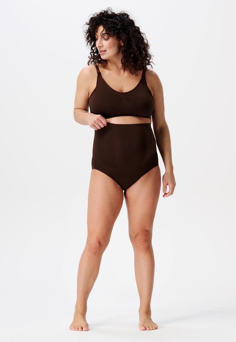 Femme aux cheveux bouclés portant un soutien-gorge seamless marron foncé et une culotte taille haute, debout pieds nus contre un fond blanc.